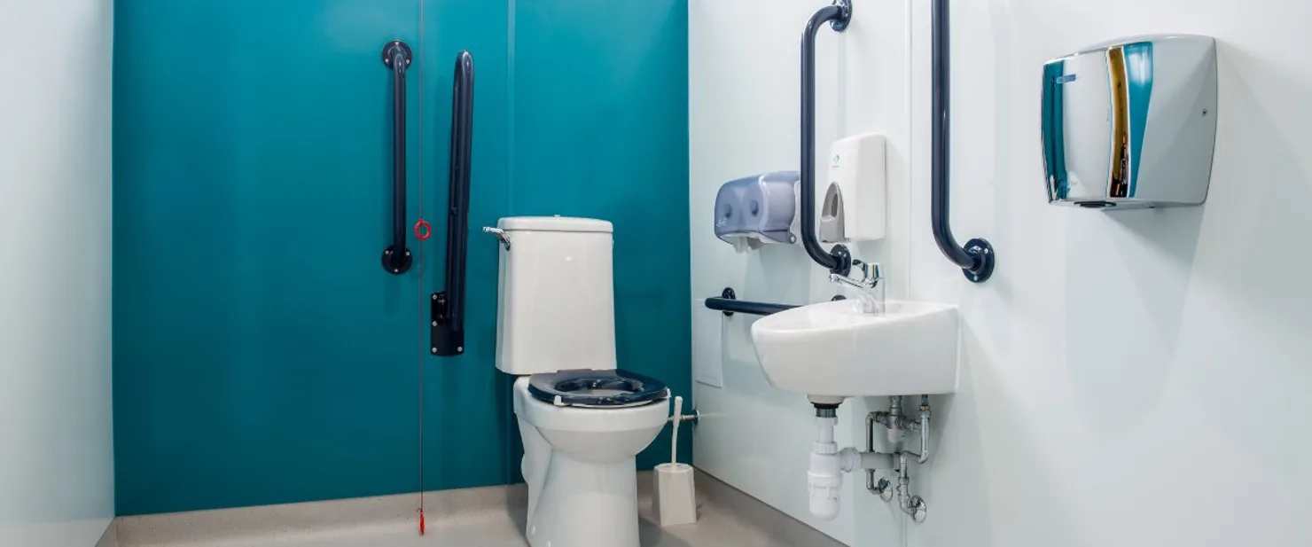 Interior of an accessible toilet showing grab rails, emergency pull cord, lowered sink, toilet paper dispenser and hand dryer